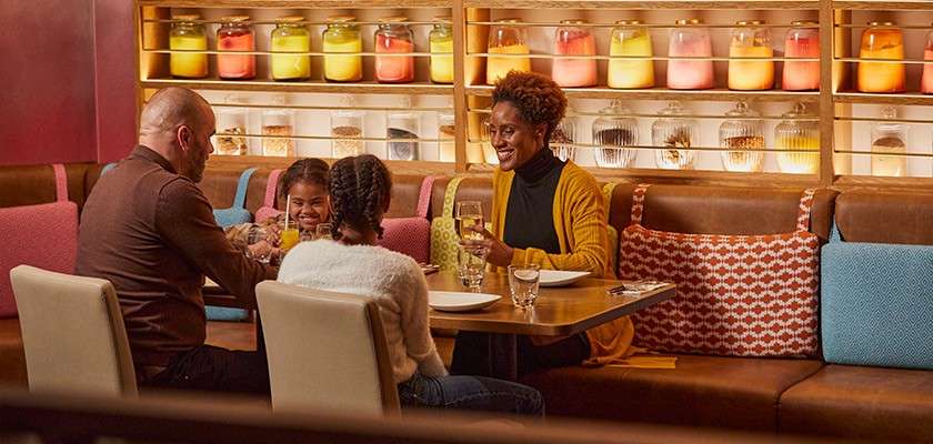 A family of four dines, smiling and talking, at a restaurant booth; plates, glasses, and silverware on the table; colorful cushions and glowing jar lights lining shelves behind.
