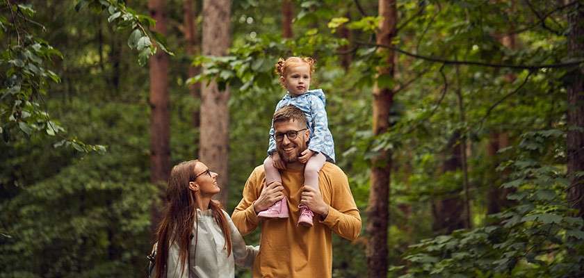 Child riding on an adult’s shoulders, another adult walking beside them, all smiling, surrounded by tall trees and dense green foliage in a forest.