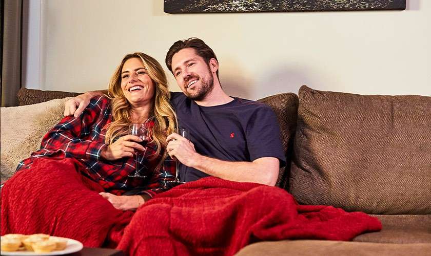 Two people cuddle on a couch, sipping wine and smiling, wrapped in a red blanket; they relax in a living room with brown cushions, a snack plate visible in foreground.