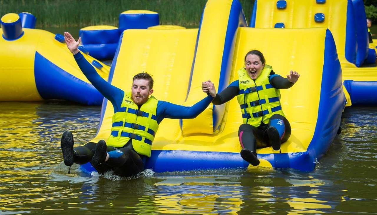 Couple sliding down Aqua Parc inflatable obstacle course on the lake.