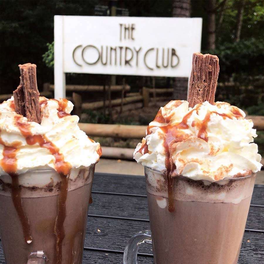 Two hot chocolates topped with whipped cream, caramel drizzle, and chocolate flakes sit on a dark outdoor table; background features a white sign reading: THE COUNTRY CLUB.