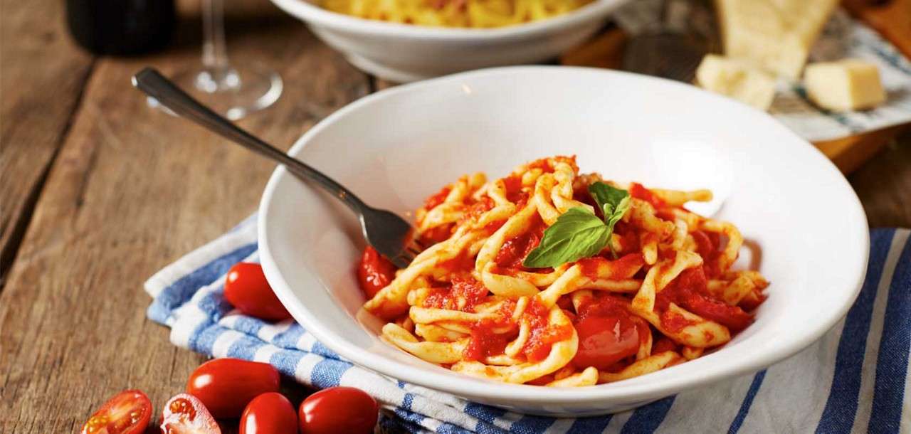 Bowl of pasta in tomato sauce rests garnished with basil. A spoon leans inside. Surrounding, cherry tomatoes, a blue-striped towel, rustic wooden table, blurred wine glass, and bread with cheese in background.