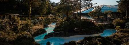 A winding lazy river snakes through landscaped rocks and pines, shimmering under sunlight; wooden fences and a glass-roofed building border the water within a tranquil, forested resort setting.