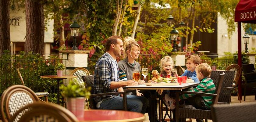 Family eats and laughs at an outdoor café table, parents and three children sharing burgers and drinks; wicker chairs, trees, lamps, and a red awning reading "BOOKIN" surround them.