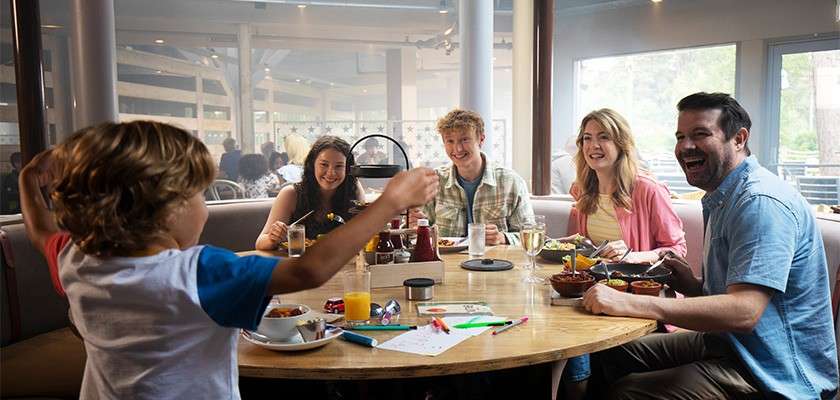 Family group dine and laugh, while a child stands with raised arms, showing something across a round table filled with bowls, drinks, and crayons, inside a bright, casual restaurant booth.