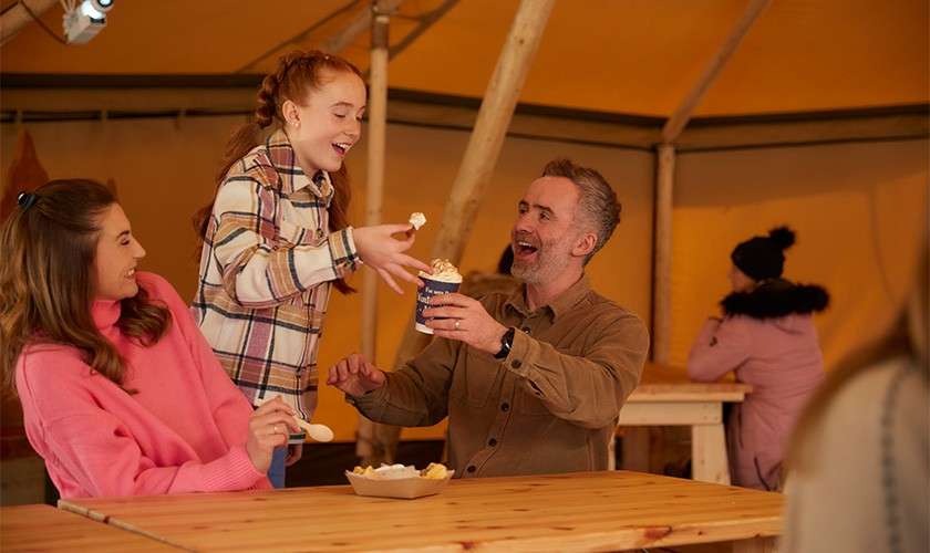 Girl hands a whipped-cream-topped drink to a smiling man while a woman watches, all seated at wooden tables inside a canvas tent; another person in a coat sits in the background.