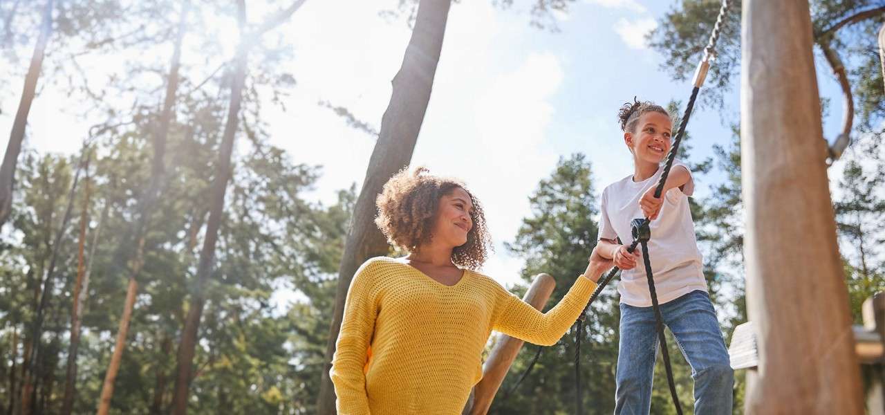 Group of bundled-up people gaze upward and smile, standing on a forest path, surrounded by tall pines, vertical light tubes on trunks, and small glowing sculptures lining the trail.