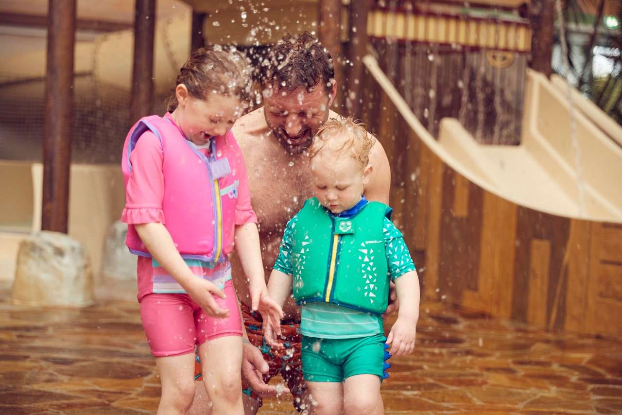 Children wearing pink and green life vests splash and laugh under falling water, while an adult kneels between them, inside an indoor water park near slides and wooden structures.