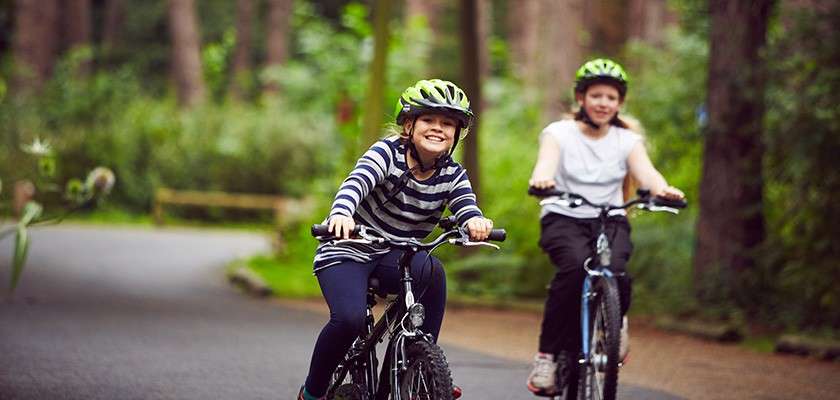 Two children on bicycles ride forward, smiling, wearing helmets; context: a winding paved path through a green forest, trees surrounding them and a softly blurred background.