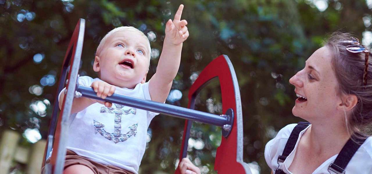 Child points upward while riding a playground rocker, gripping a metal bar; adult smiles beside them; leafy park background with dappled light.