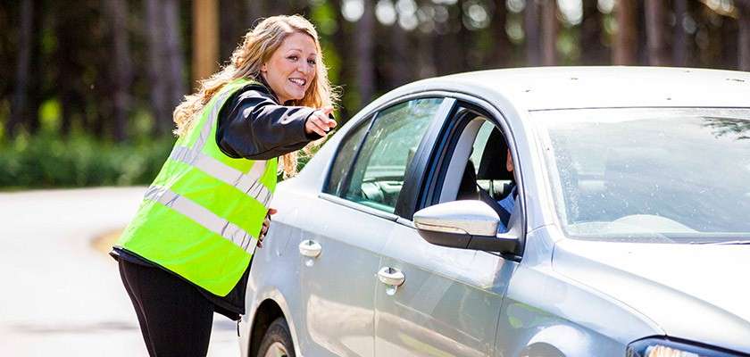 Traffic marshal in high-visibility vest leans toward a silver car, pointing to direct the driver. Scene occurs on a sunny roadside with blurred trees in the background.