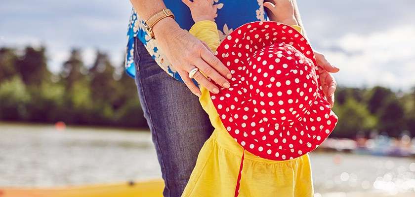 Child in a red polka-dot sunhat holds an adult’s hands, looking up. They stand lakeside on a sunny day, with water, distant trees, and boats blurred in the background.
