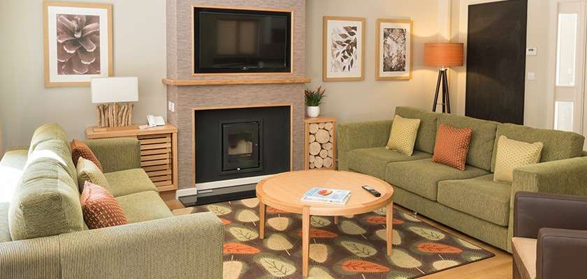 Green sofas frame a round coffee table holding magazine and remote, while a wall-mounted TV and fireplace anchor a cozy room with leaf-pattern rug, botanical artwork, and stacked firewood.