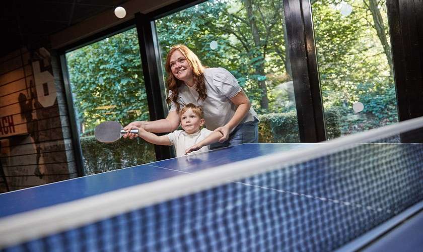 Adult and child swing a table-tennis paddle together, returning a ball over a blue ping-pong table. They stand indoors by tall windows overlooking green trees; wall art shows the number “6”.