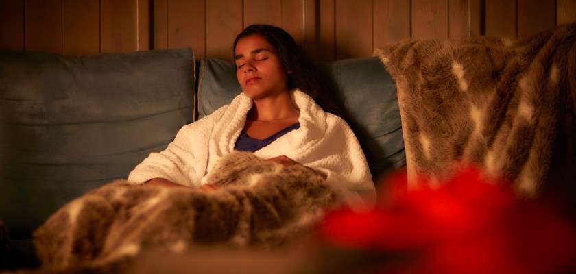 Person rests with eyes closed, wrapped in a white robe and furry blanket, reclining on a cushioned sofa in warm, dim lighting against wood-paneled walls, creating a cozy indoor scene.