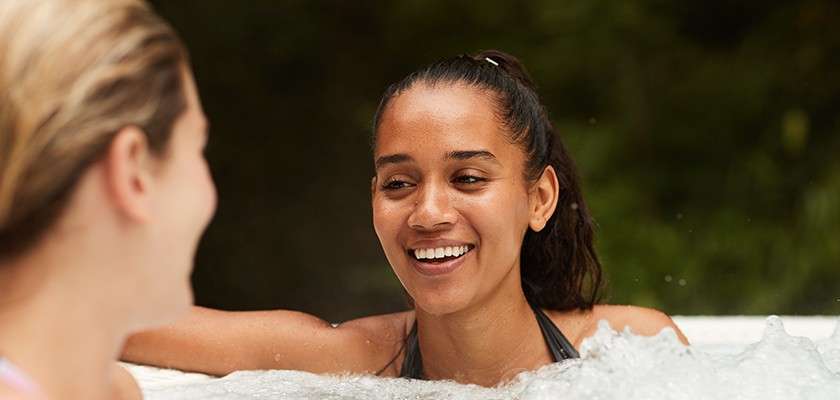Two people sit in a bubbling hot tub, one smiling and speaking while resting an arm on the edge; the other listens. Background shows greenery, suggesting an outdoor, relaxed setting.