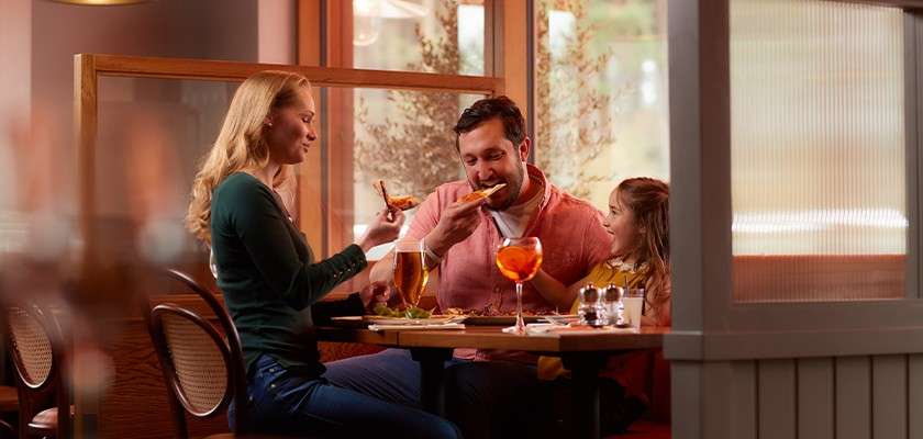 Family eats pizza at a booth in a cozy restaurant. Adults hold beer and an orange cocktail while a child smiles beside them amid warm lighting and wooden partitions.