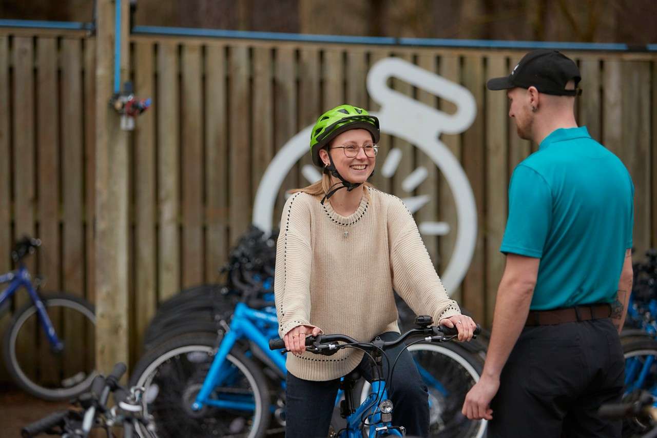 Cyclist smiles while holding handlebars, talking to a staff member; rental bikes line a rack behind them in an outdoor bike station with a wooden fence and a large stopwatch graphic.
