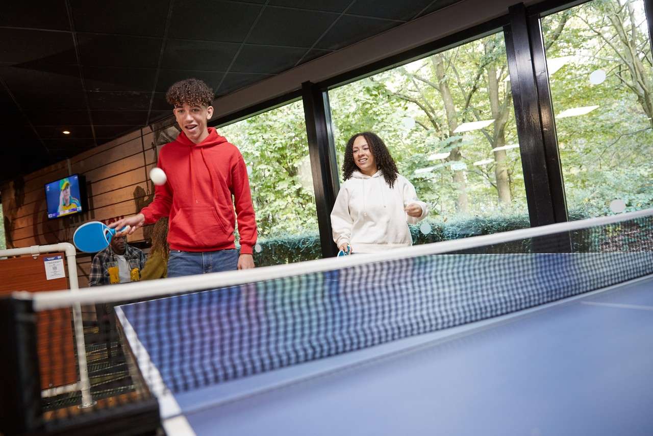 Two people play table tennis, paddles up as a white ball hovers over the net, inside a lounge with big windows to leafy trees, a wall-mounted TV, and a seated onlooker.