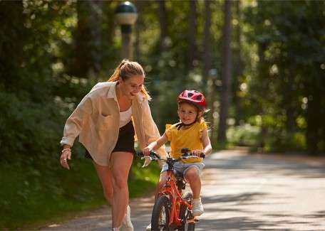 Child rides a small bike; adult jogs beside, guiding a hand near the seat; in a leafy park path with dappled sunlight, the child wears a red helmet and training wheels.