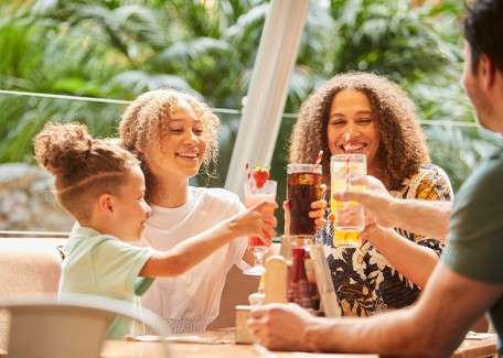 Four people raise drinks, clinking glasses and smiling. They sit around a wooden table with condiments and water glasses, in a sunlit outdoor café surrounded by lush green plants.