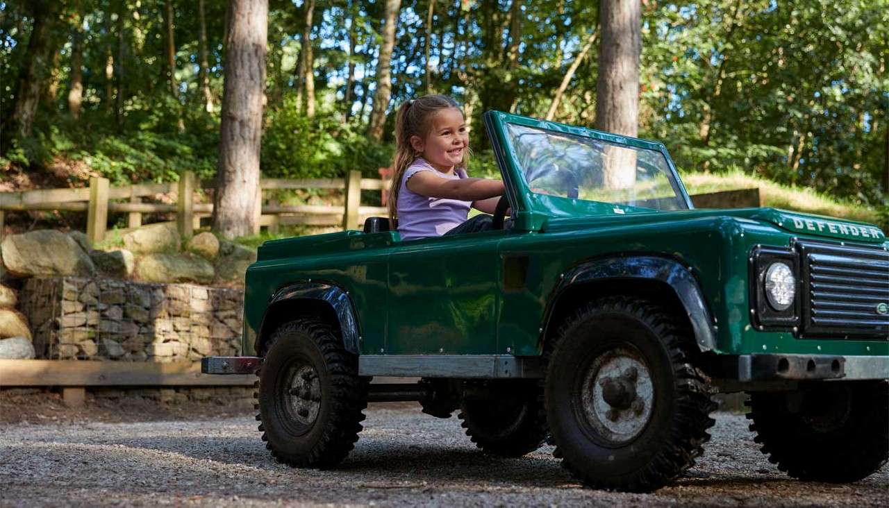 Child in a small green off-road toy car steers and smiles, driving on a gravel path; surrounding trees, wooden fence, and stone wall in sunlight. Visible text: DEFENDER.