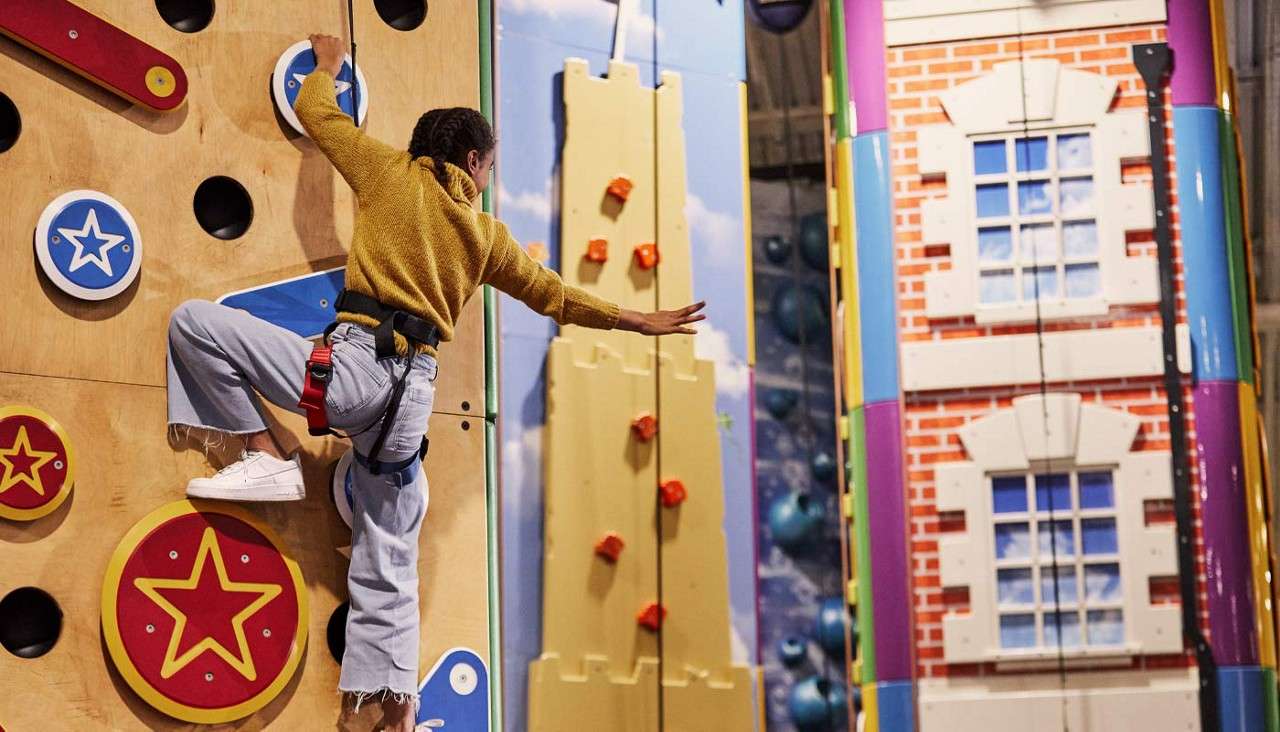 Person climbs a colorful indoor climbing wall, reaching sideways while harnessed. Surroundings show playful murals and themed climbing panels, including star-decorated holds and building facade graphics in a recreation facility.