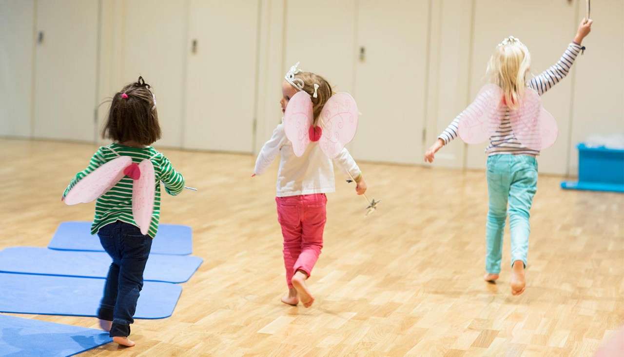 Three children wearing fairy wings run barefoot, some waving wands, across a wooden-floored room with blue mats and closed doors.