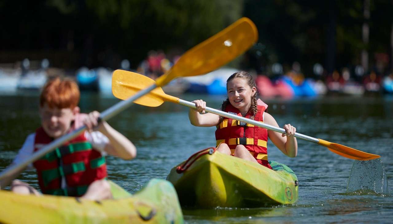 Two children in red life jackets paddle bright green kayaks, splashing water, on a calm lake; blurred boats and trees fill the sunny background.