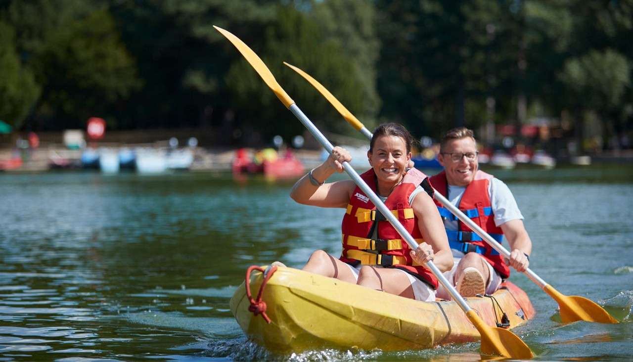 Two people paddle a yellow tandem kayak, wearing red life vests, smiling. On a calm lake, trees and moored boats blur in the background. Text: JOBE.