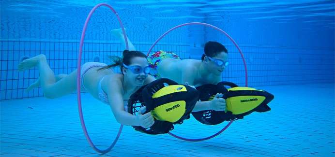 Two swimmers use yellow handheld underwater scooters, gliding forward through pink hoops in a tiled pool. Text on scooters: SEADOO, SEADOO.