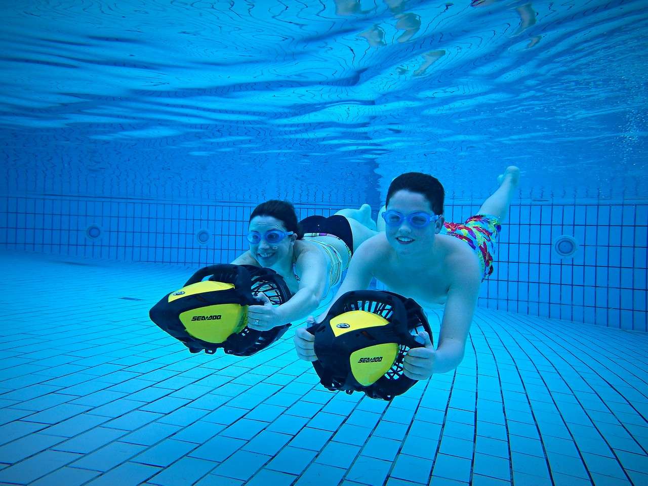 Two swimmers use yellow handheld underwater scooters, gliding forward through pink hoops in a tiled pool. Text on scooters: SEADOO, SEADOO.