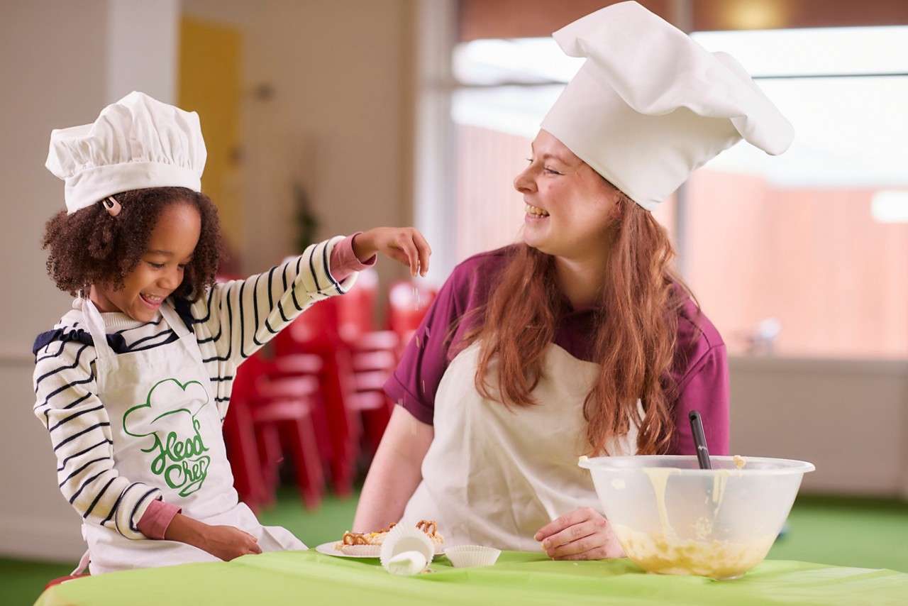 Child sprinkles ingredients while a smiling chef supervises, both wearing white hats and aprons, in a bright kitchen classroom with blurred red chairs and utensils in the background.