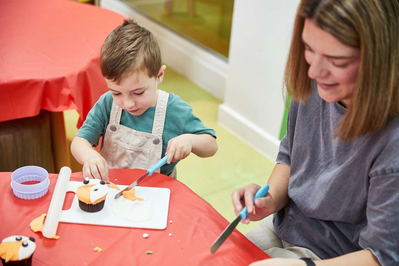 Child wearing safety goggles watches a small homemade volcano bubble with pink foam in a yellow bin, while an adult supervises in a classroom with green walls and posters.