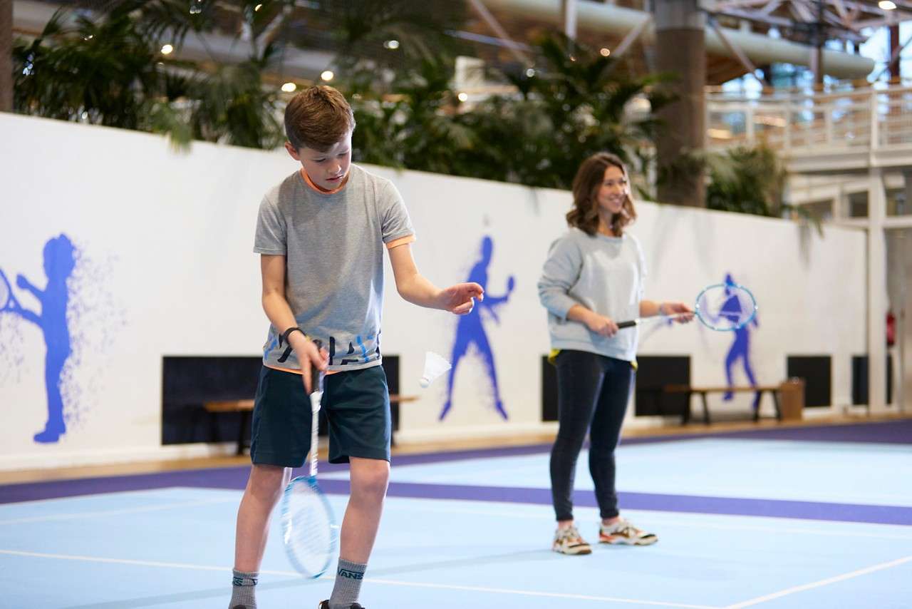 Smiling child runs, clutching a blue ball, in an indoor gym. Yellow inflatable barriers edge the court while colorful walls and another child in back suggest a playful sports session.