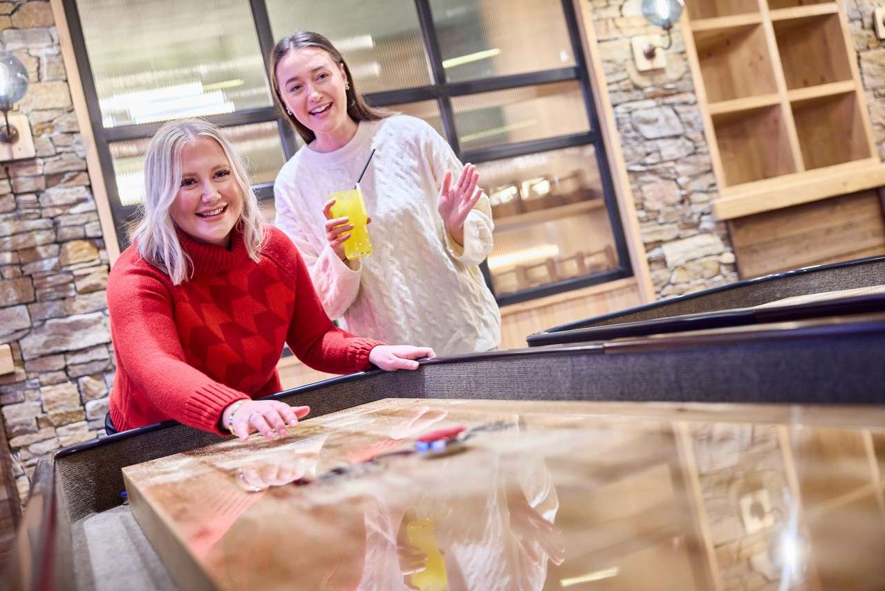 Two people play; one slides a shuffleboard puck while the other watches, holding a yellow drink. A cozy indoor lounge surrounds them with stone walls, wood shelves, and large windows.
