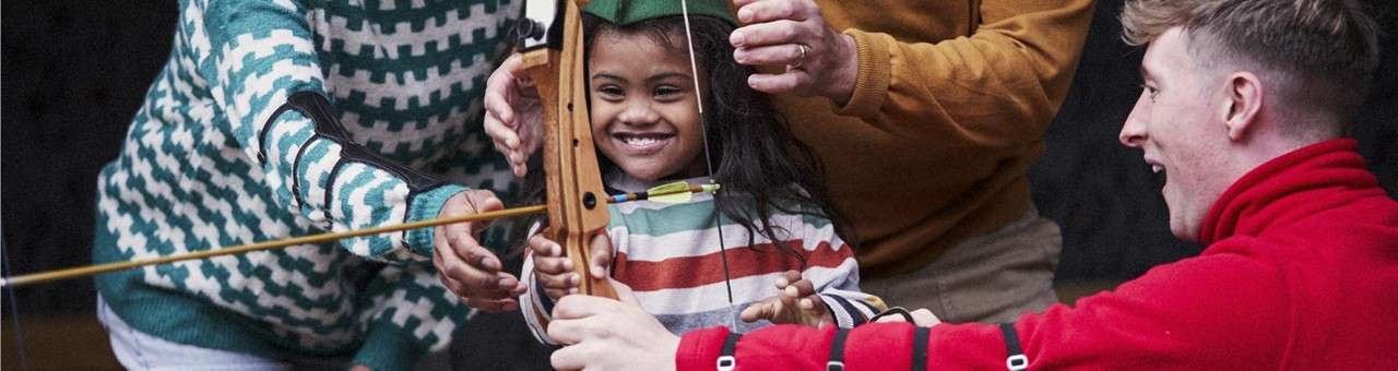 Child holds a wooden bow, guided by several adults’ hands, preparing to shoot an arrow; people smile and wear casual, warm clothing, suggesting an archery lesson in a recreational setting.