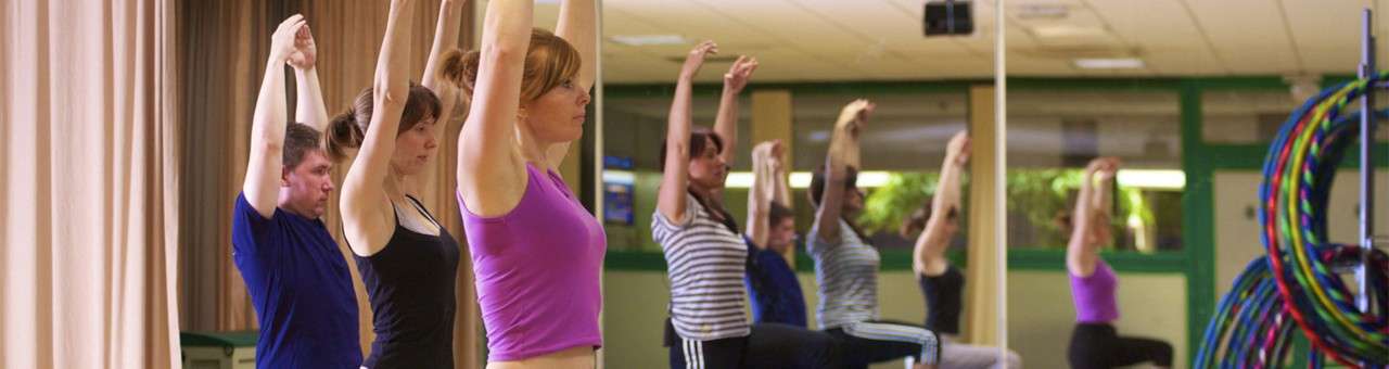 Class participants raise arms while holding lunging poses, aligned in a row. They exercise in a mirrored gym studio with green walls, fluorescent lighting, and stacked colorful hula hoops.
