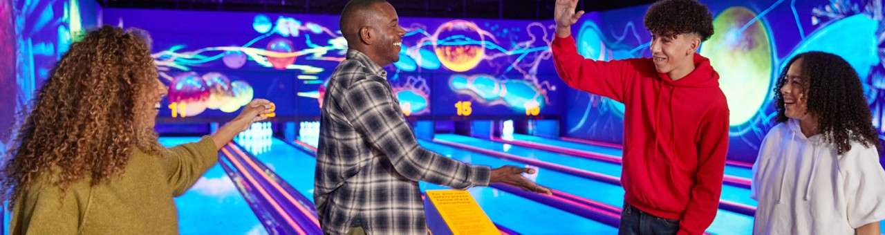 Four people high-five and smile, celebrating a bowling turn, in a neon-lit alley with colorful cosmic murals and glowing lanes, with "15" displayed on the back wall above pins.