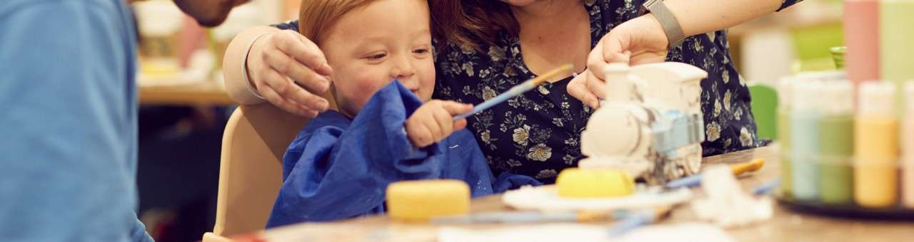 Child paints a small ceramic train while two adults guide their hands, using brushes and bright paints; jars, sponges, and art supplies sit on a table in a busy studio.