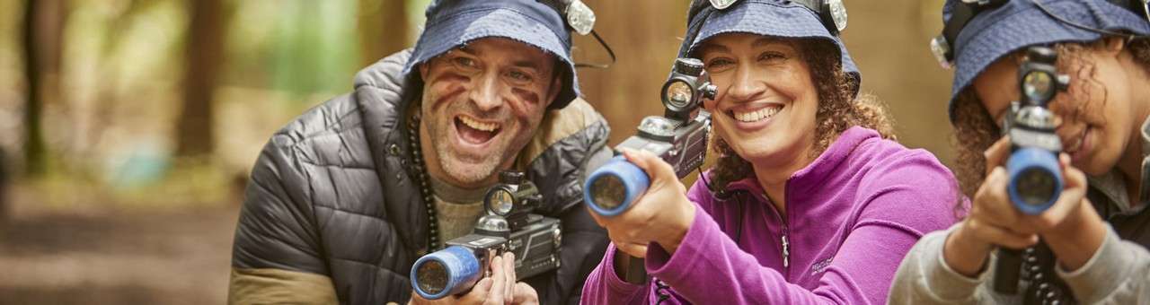 Three smiling people aim laser-tag guns forward. Wearing bucket hats with headlamps and face paint, they play together in a wooded outdoor area with blurred trees and warm light.