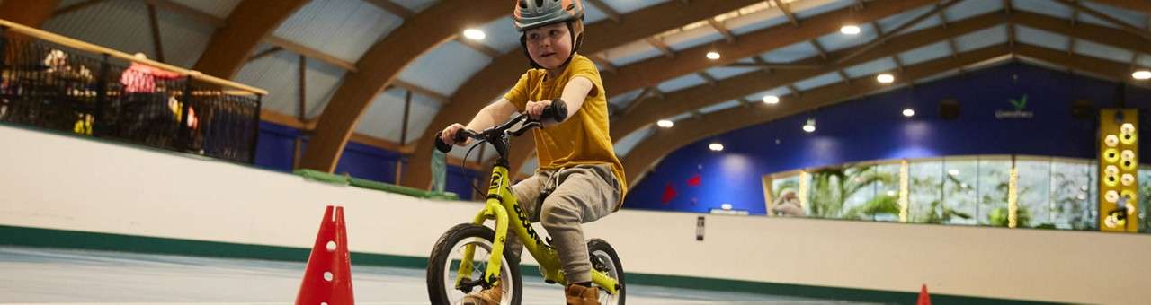 Child on a balance bike rides around red cones, wearing a helmet, inside a spacious indoor track with arched wooden beams, bright lights, and onlookers behind a railing.