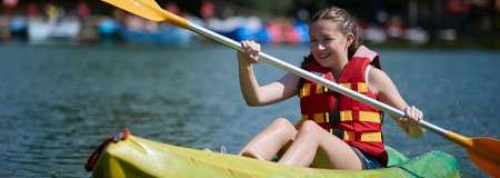 Child paddles a yellow kayak, smiling and rowing with a double-bladed paddle, on calm lake water; blurred boats and trees line the distant shoreline.