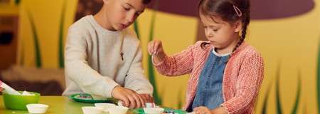 Two young children assemble crafts, dipping tools into small bowls and arranging pieces on a tray, at a green table in a colorful playroom with yellow walls and grass-themed decor.