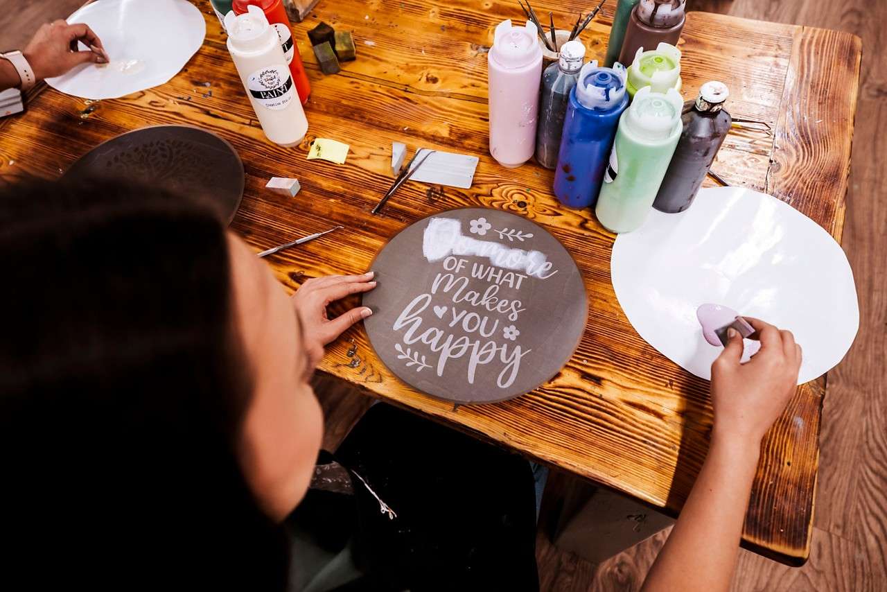 Hands stencil lettering onto a round gray board on a wooden craft table with paint bottles and tools. Text: “more OF WHAT MAKES YOU happy.” Bottle label: “PAINT.”