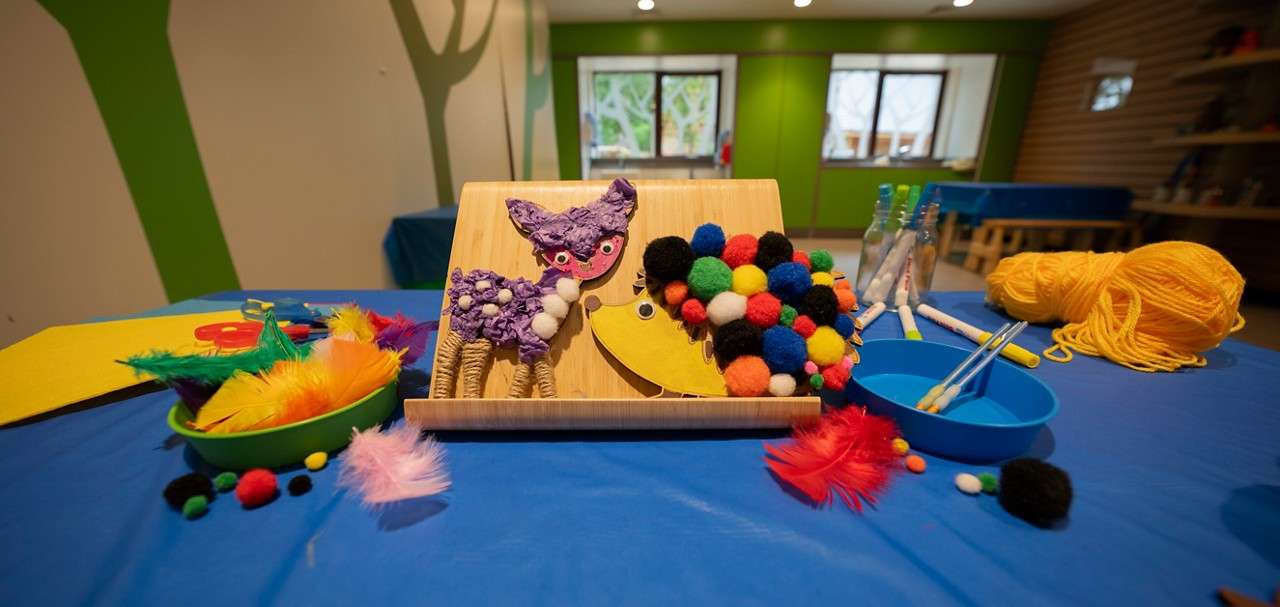 Children’s craft display shows decorated animal cutouts resting on a wooden stand, surrounded by bowls of pom-poms, feathers, markers, and yellow yarn on a blue table in a bright classroom.
