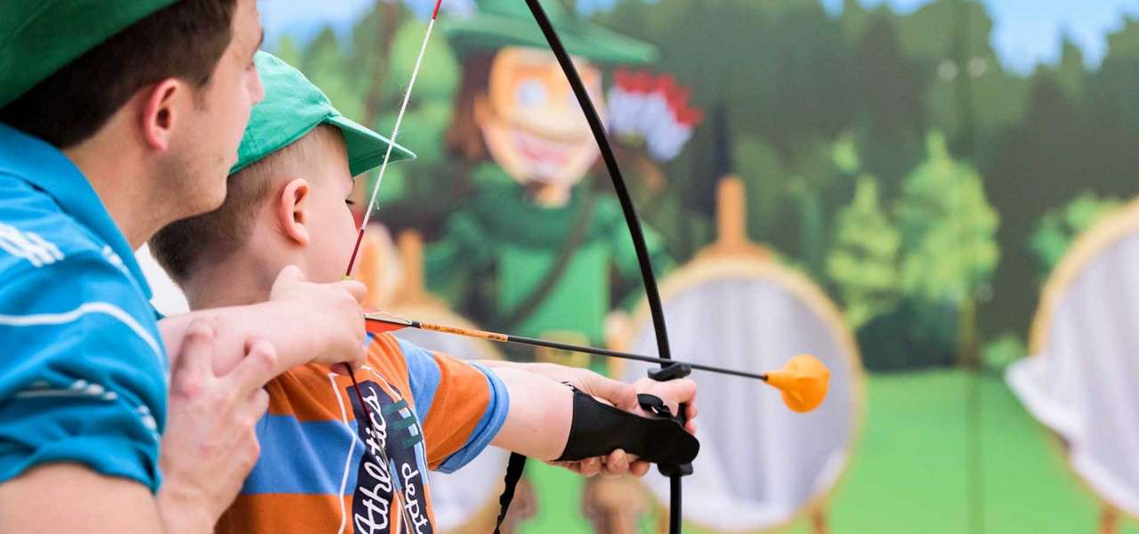 Child archer aims a suction-cup arrow while an adult guides their arms, in a playful archery range with cartoon forest backdrop and round targets. Shirt text reads: "Athletics".