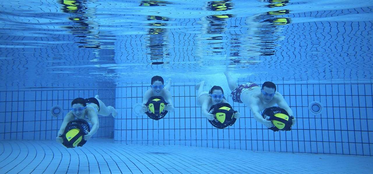 Five swimmers hold handheld underwater scooters, gliding forward in formation; context: a clear blue tiled swimming pool with grid-patterned walls and floor, bubbles and reflections visible near the surface.