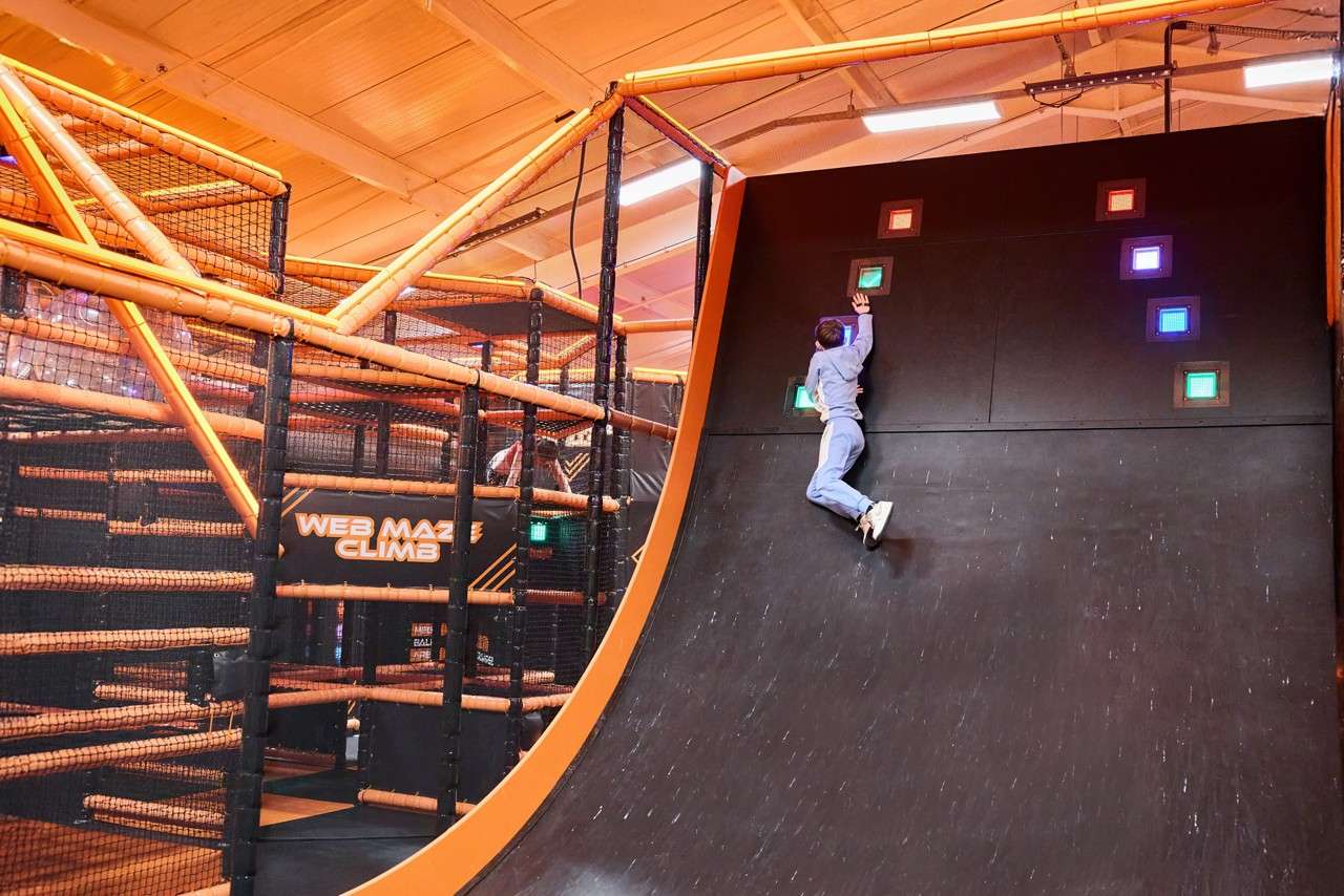 Person clings mid-climb on a steep black curved wall, reaching for lit square targets; in an indoor orange-framed obstacle course with netted platforms and a sign reading WEB MAZE CLIMB.