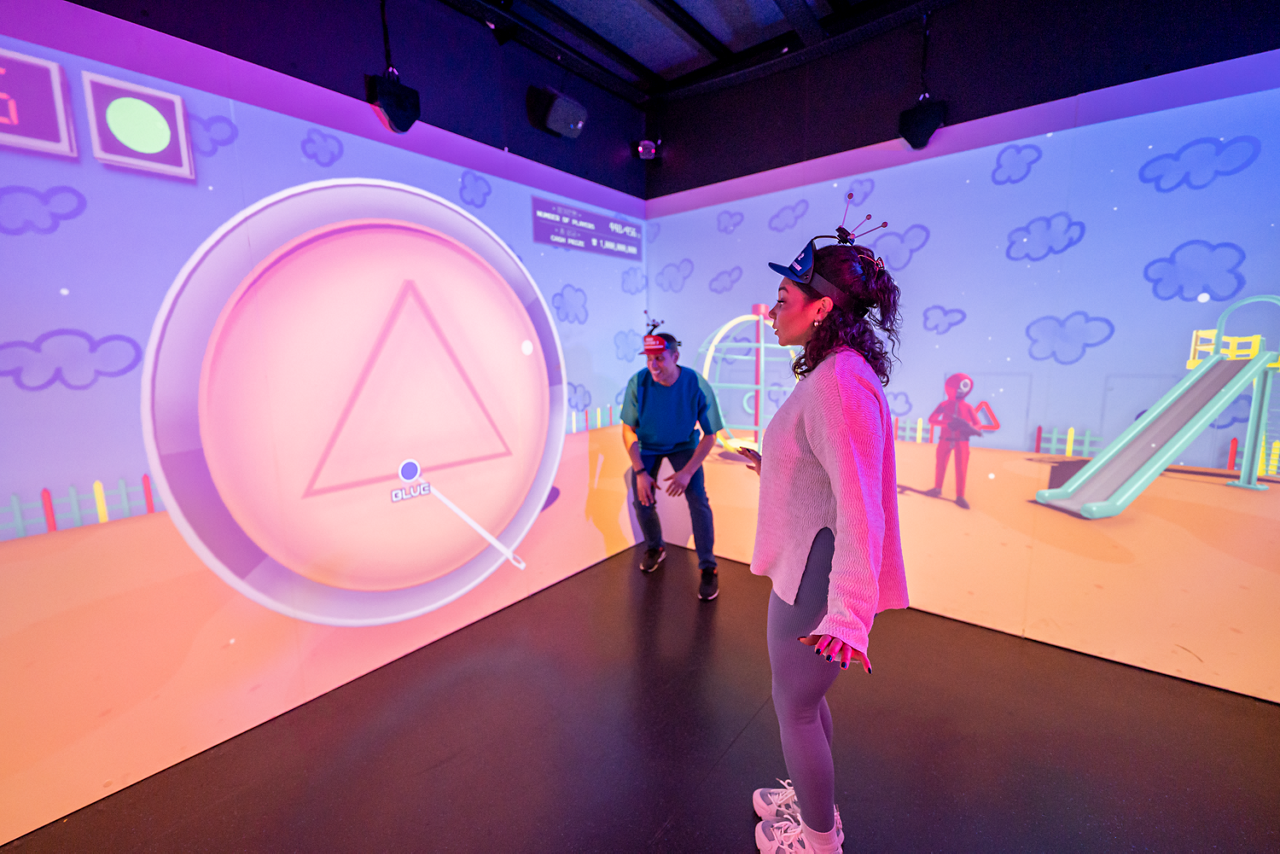 Two players wearing sensor caps gesture, interacting with a large projected dial showing a triangle and cursor labeled “BLUE,” inside an immersive room featuring cartoon playground walls and a slide.